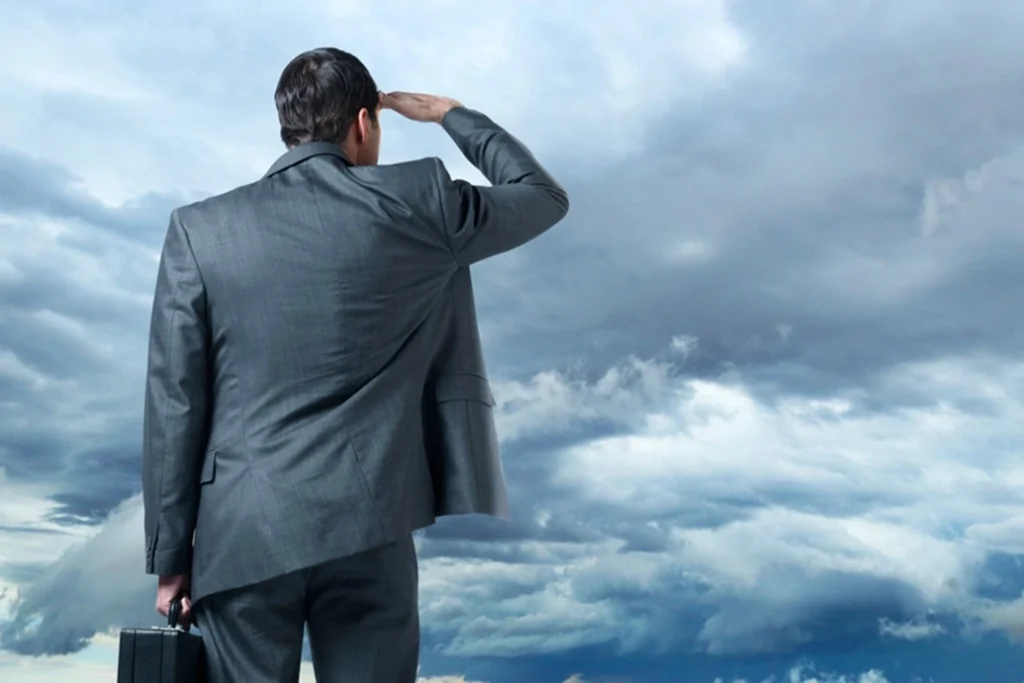 A man in a gray suit holding a briefcase stands with his back to the camera, shielding his eyes as he looks towards a cloudy sky.