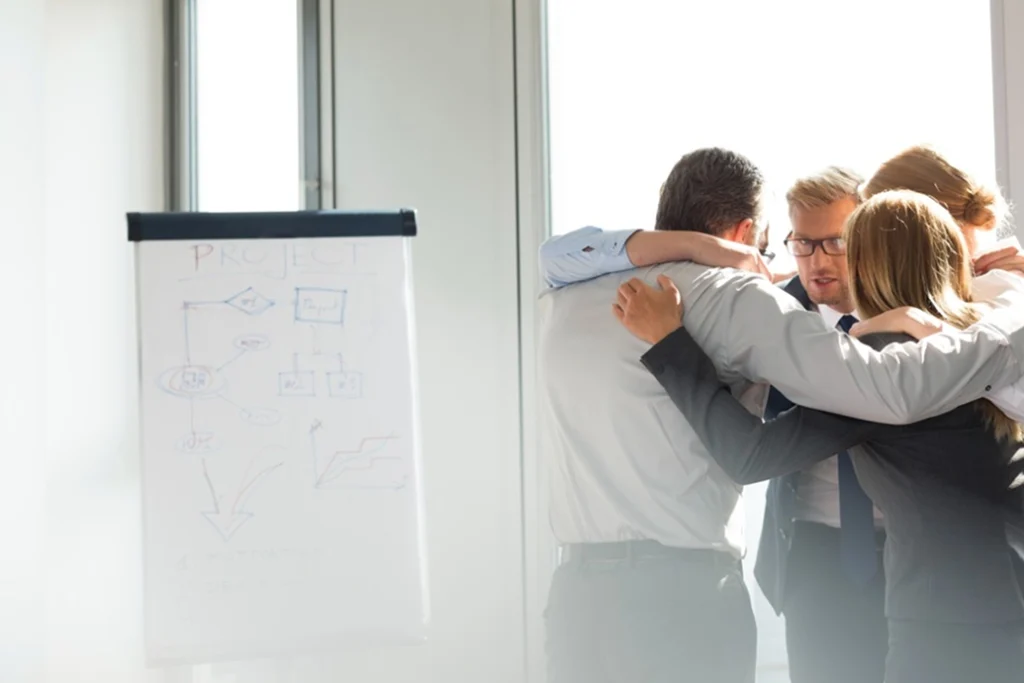 Four business professionals stand in a close huddle, smiling and embracing in a bright office. To the left, a flip chart displays diagrams and the word “PROJECT” at the top. Sunlight streams through large windows.
