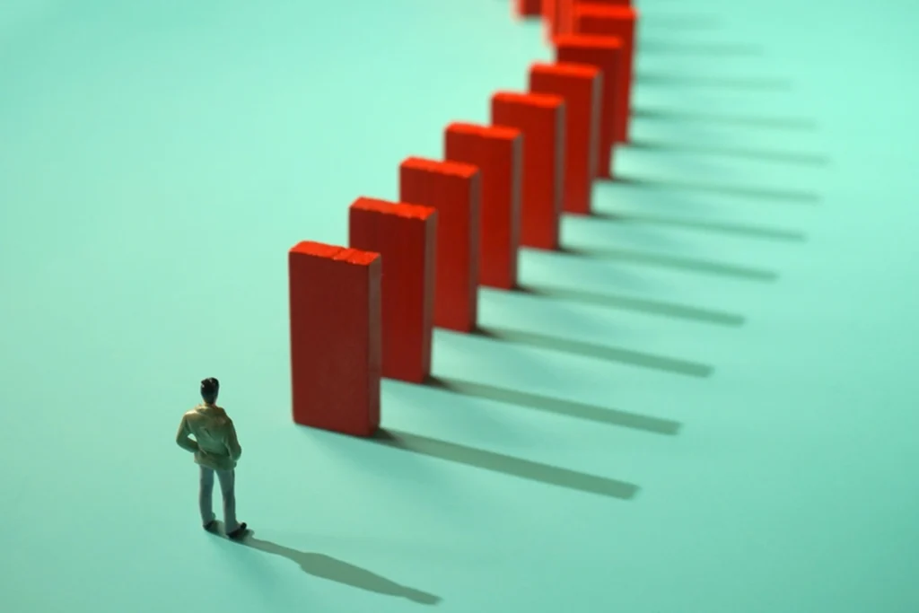 A small figurine of a person stands facing a curved line of upright red dominoes casting long shadows on a teal surface, suggesting anticipation or cause and effect.