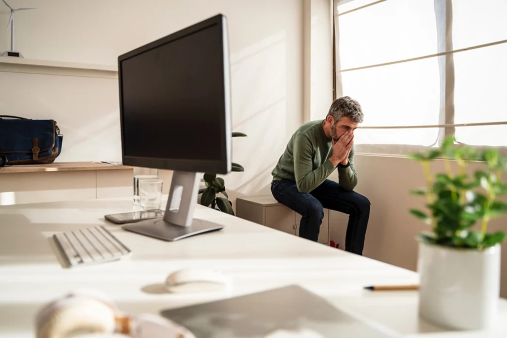 A man sits alone in an office, covering his face with his hands in distress. A computer and office supplies are on a desk in the foreground, with sunlight streaming through the window.