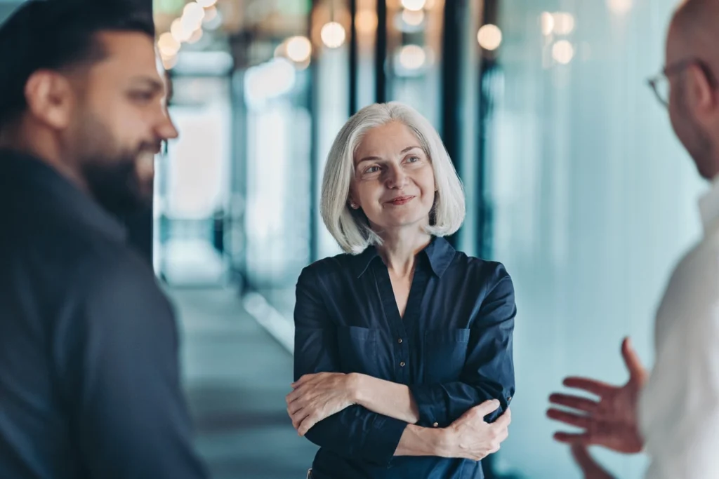 A woman with gray hair and a navy shirt stands with arms crossed, smiling as she listens to two men talking in a modern office hallway with glass walls and warm lighting.