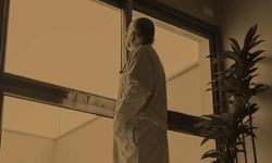 A person wearing a lab coat stands indoors, looking out a large window; a tall plant is visible on the right side.
