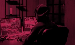 A person in a hoodie sits at a desk using two computer monitors displaying various screens, in a dimly lit room with a red tint.
